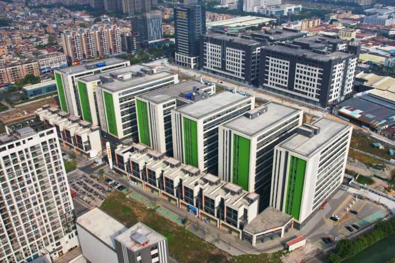 Aerial view of a modern urban area with several large, multi-story buildings featuring green accents, surrounded by other office buildings, roads, parked cars, and dense cityscape in the background.