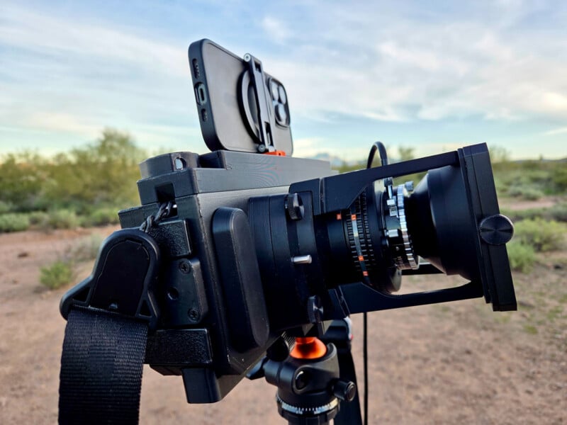 A close-up of a camera mounted on a tripod in an outdoor setting, with a smartphone attached on top. Desert vegetation and a cloudy sky are visible in the blurred background.