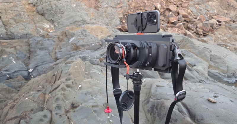 A large-format film camera with a smartphone attached on top is mounted on a tripod on rocky terrain, showing a blend of modern and traditional photography equipment.