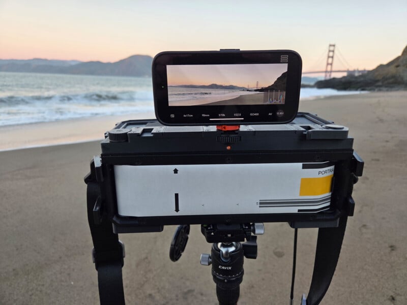 A panoramic camera on a tripod is set up on a sandy beach, capturing the Golden Gate Bridge at sunset. The bridge and ocean appear on the camera's digital screen. Mountains and calm water are in the background.