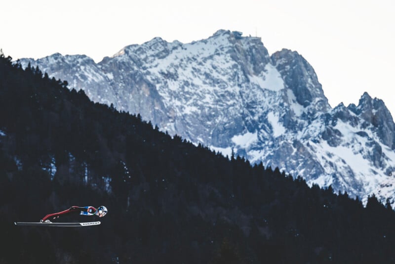 A ski jumper in mid-air soars against a backdrop of tall, snow-capped mountains and a dark forested hillside. The athlete is wearing a helmet and a suit, with skis extended forward.