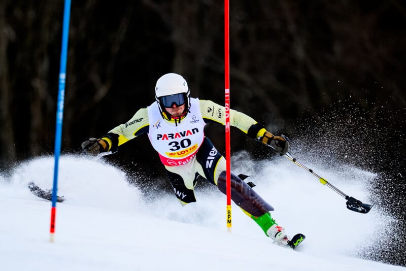 A skier wearing a helmet, goggles, and race bib number 30 makes a sharp turn during a slalom race, kicking up snow while maneuvering between red and blue slalom poles on a snowy slope.