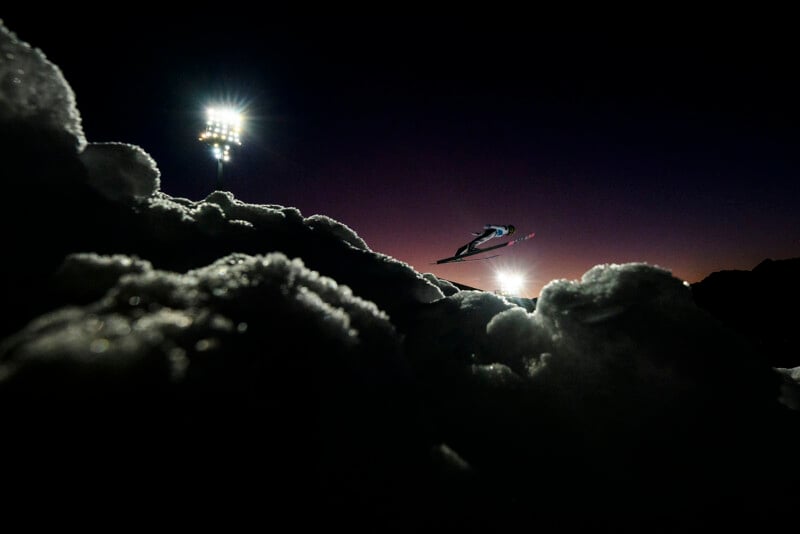 A ski jumper is captured mid-air against a dark night sky, with bright stadium lights illuminating snow in the foreground. The scene is dramatic and dynamic, highlighting the skier’s silhouette and motion.