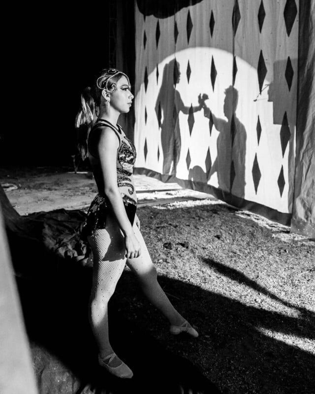 A circus performer in costume stands backstage, looking at a stage with diamond-patterned curtains and a spotlight casting her shadow. The scene is in black and white.