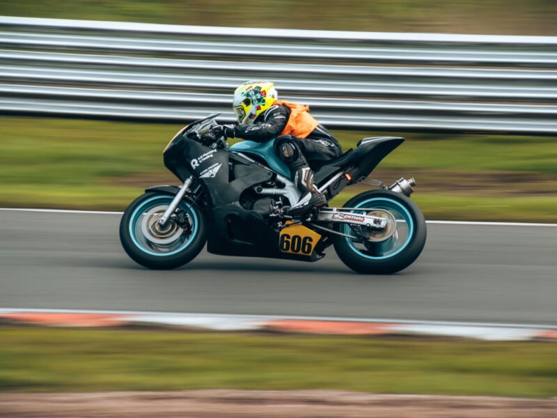 A motorcyclist wearing a yellow helmet and orange vest speeds on a black racing motorcycle with the number 606 on a curving racetrack, the background blurred to show motion.
