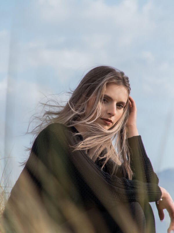 A young woman with long, light brown hair sits outdoors in front of a cloudy sky, wearing a black sweater and looking thoughtfully at the camera, with grass softly blurred in the foreground.
