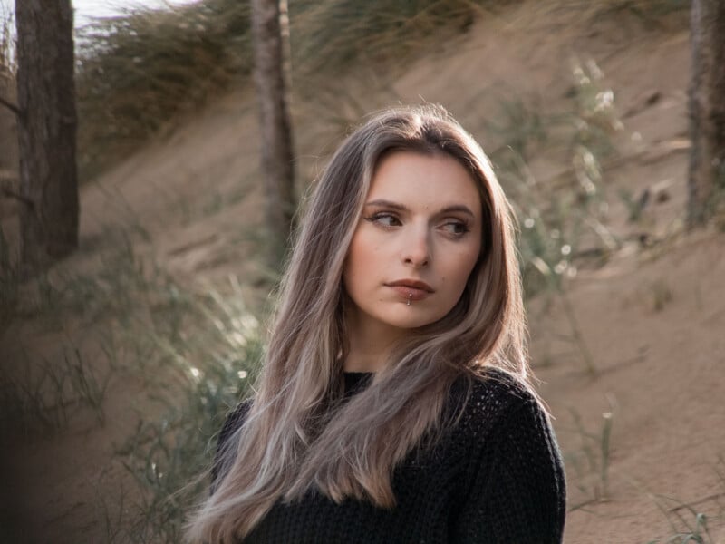 A young woman with long, light brown hair and a black sweater stands outdoors on sandy terrain with grasses and trees, looking to her left with a thoughtful expression.
