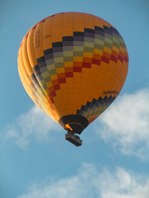 A colorful hot air balloon with orange, yellow, black, and gray patterns floats in the sky above scattered clouds.