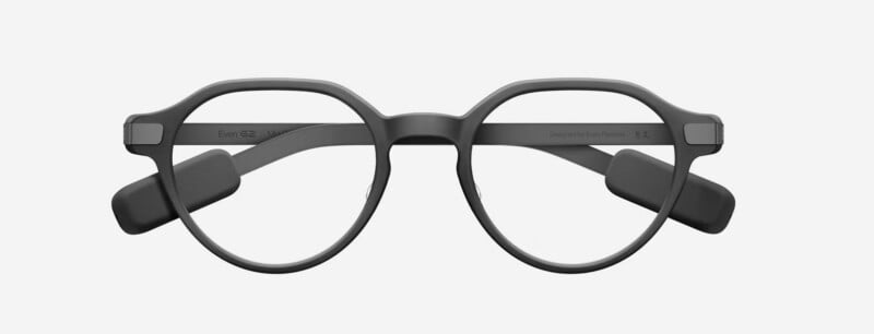 A pair of round, matte black smart glasses viewed from the front, featuring thick frames and arms with rectangular modules attached near the hinges.