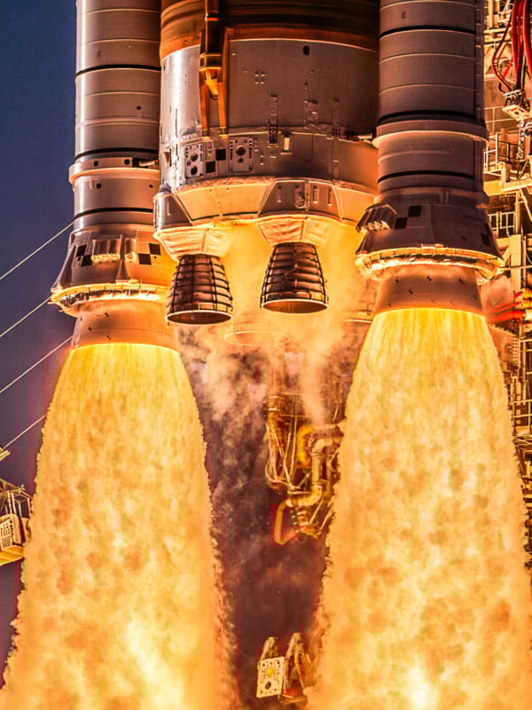 Close-up view of a rocket launch, showing intense orange flames and smoke erupting from the engines as the spacecraft lifts off, with structural details and equipment visible around the base.