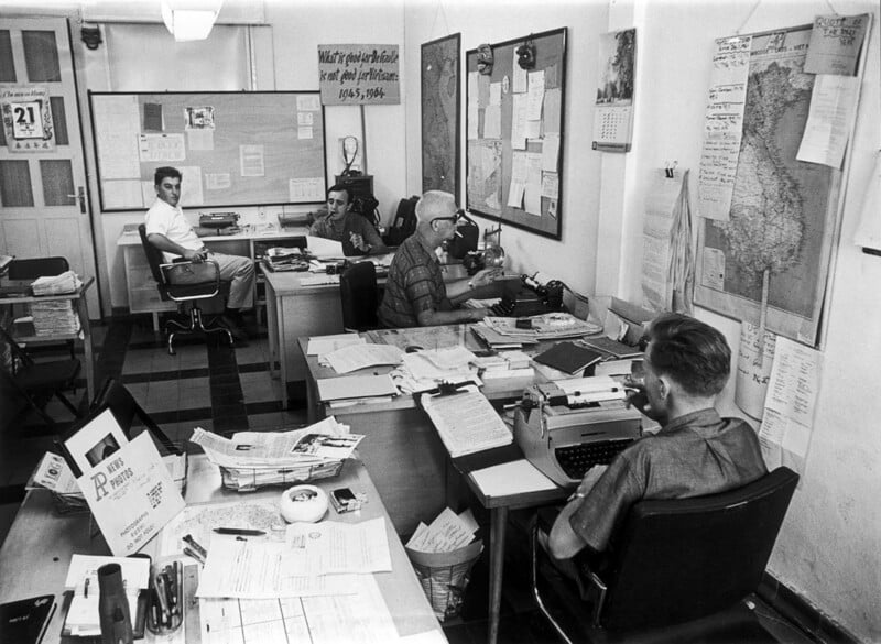 Four men work at desks in a cluttered office filled with papers, maps, and bulletin boards. One types on a typewriter while others write or talk. The room has a busy and organized atmosphere.