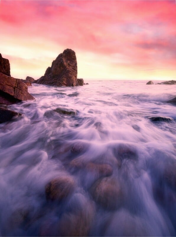 Rocky shoreline with waves washing over smooth stones at sunset, pink and orange clouds fill the sky, and a large rock formation is visible in the distance.