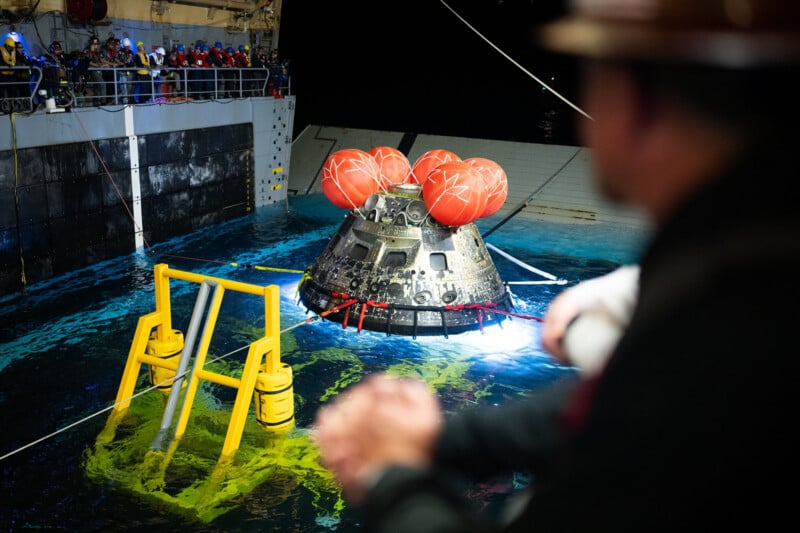 A spacecraft capsule with red flotation devices floats in the water near a recovery ship at night, as people onboard observe the retrieval operation.