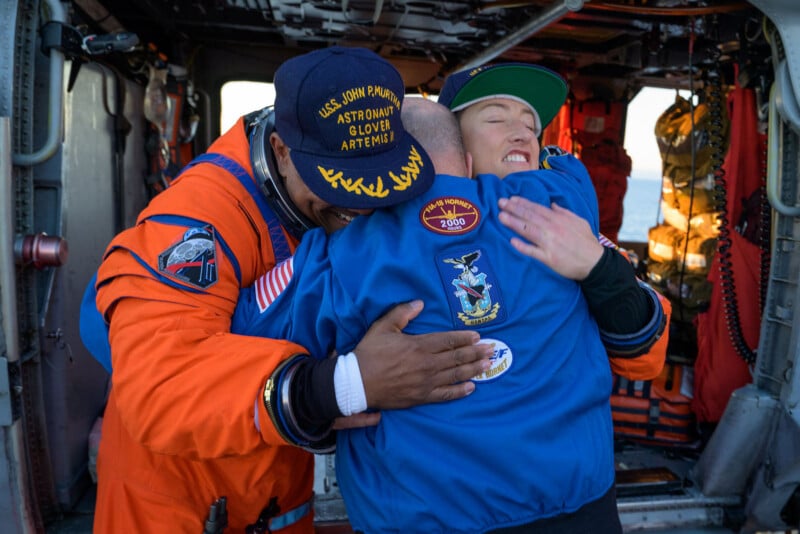 Two astronauts in orange and blue NASA flight suits embrace inside a helicopter, smiling and celebrating. Flight gear and equipment are visible in the background, suggesting a recent mission or rescue.