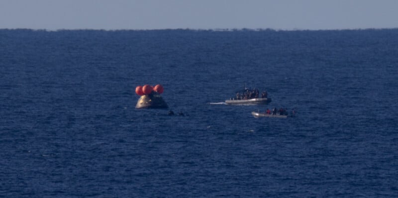 A spacecraft with orange flotation devices floats on the ocean as three boats filled with people approach it. The horizon is visible in the background under a clear sky.