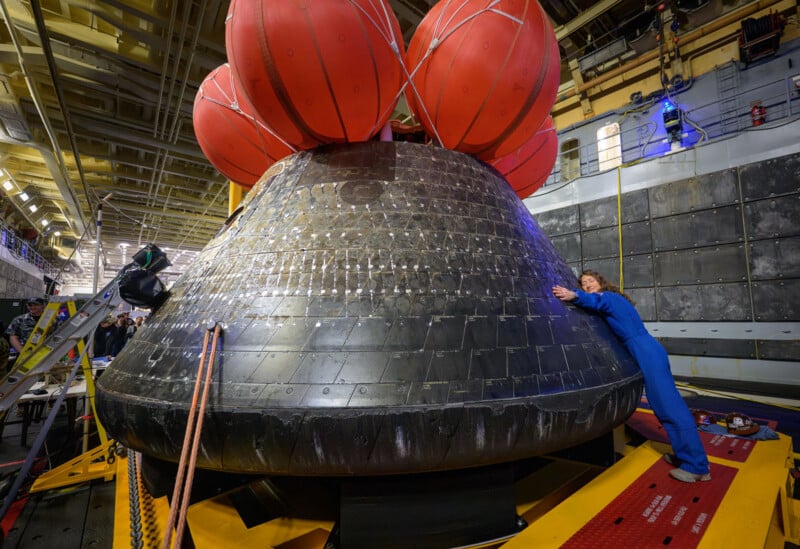 A person in a blue jumpsuit hugs a large, black, cone-shaped spacecraft capsule with red flotation devices attached, inside a brightly lit industrial facility.
