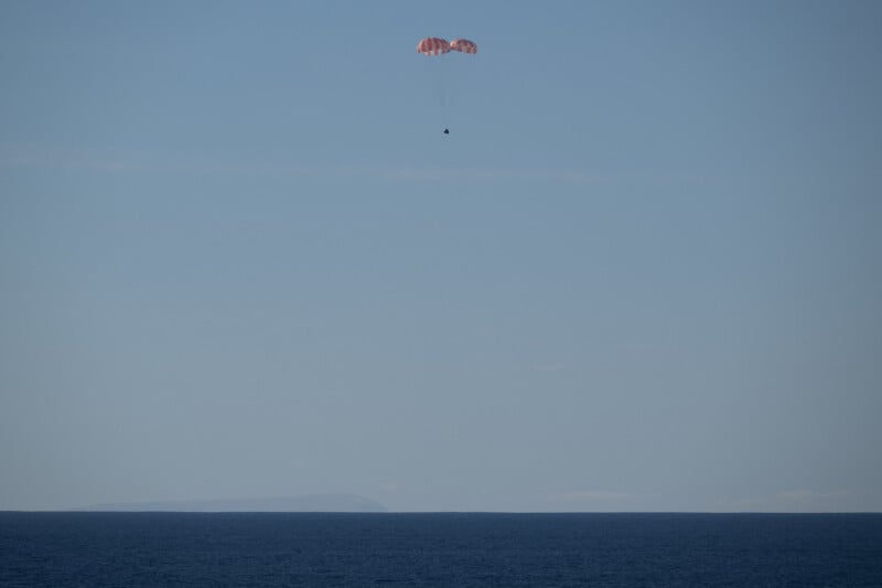 A small capsule with a red and white parachute descends toward a calm, blue ocean under a clear sky, with a faint outline of land visible on the horizon.
