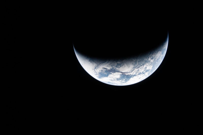 A crescent-shaped Earth is seen from space against a black background, with part of the planet illuminated and the rest in shadow, revealing blue oceans and white cloud patterns.