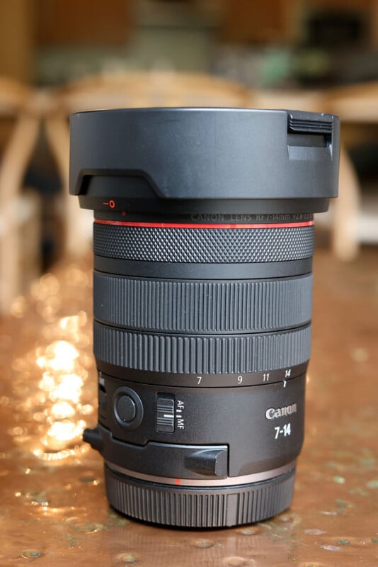 A Canon RF 14-35mm f/4 L IS USM camera lens stands upright on a brown table, with a blurred background of chairs and a room interior.