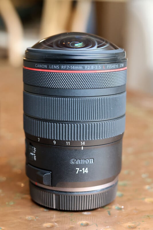 A Canon RF 7-14mm f/2.8-3.5 L fisheye STM lens is standing upright on a brown surface, showing its wide front element and detailed lens markings.