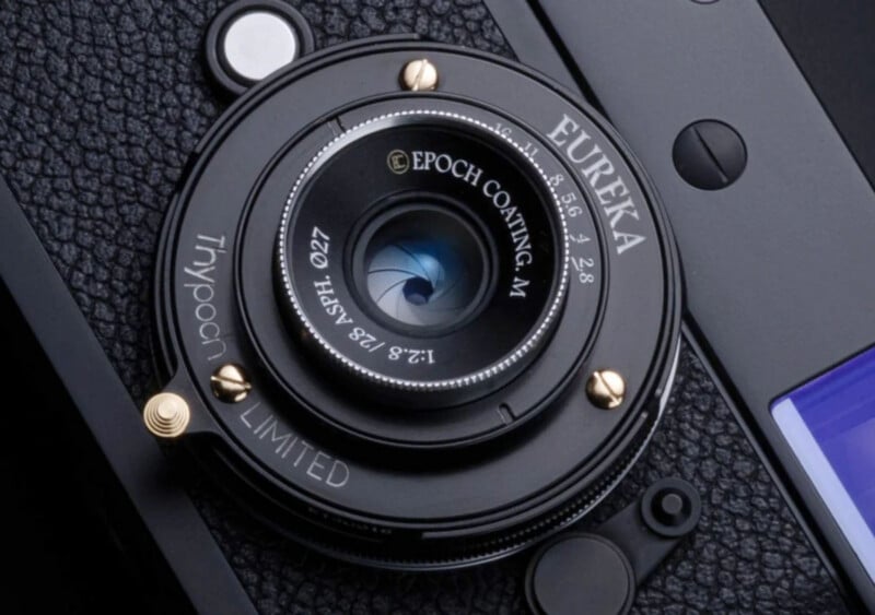 Close-up of a camera lens labeled "Eureka" with text "Epoch Coating-M," "1:2.8 f=35mm Ø27," and "Tinypoch LIMITED" visible. The camera body has a textured black surface and metal accents.