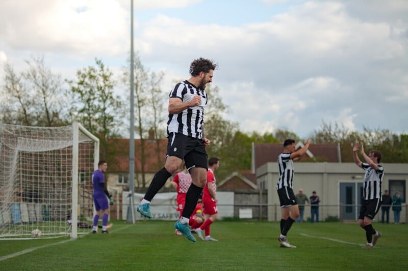 A soccer player in a black-and-white striped jersey jumps in celebration near the goal, while teammates cheer and opposing players in red stand nearby on a grassy field.
