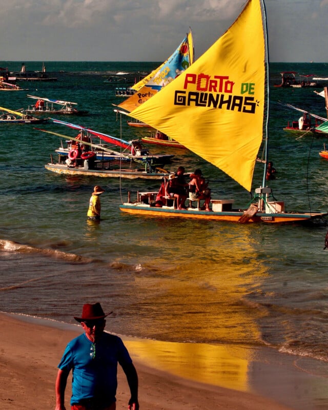 Several colorful boats with large sails float near the shore, one with a bright yellow sail reading "Porto de Galinhas." People are on the boats and beach, with a man in a blue shirt and hat walking in the foreground.