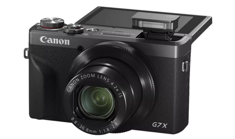 A Canon G7 X digital camera with its screen flipped up and the flash popped up, showing the zoom lens and various control dials.