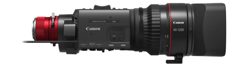 Side view of a Canon 40-1200mm broadcast camera lens with a red mount on the left, black body, control switches, and a large hood on the right.