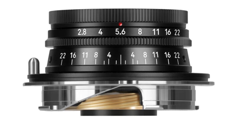 A close-up view of a black camera lens with white aperture and distance markings, set against a white background; part of the lens body is cut away to reveal internal mechanical components.