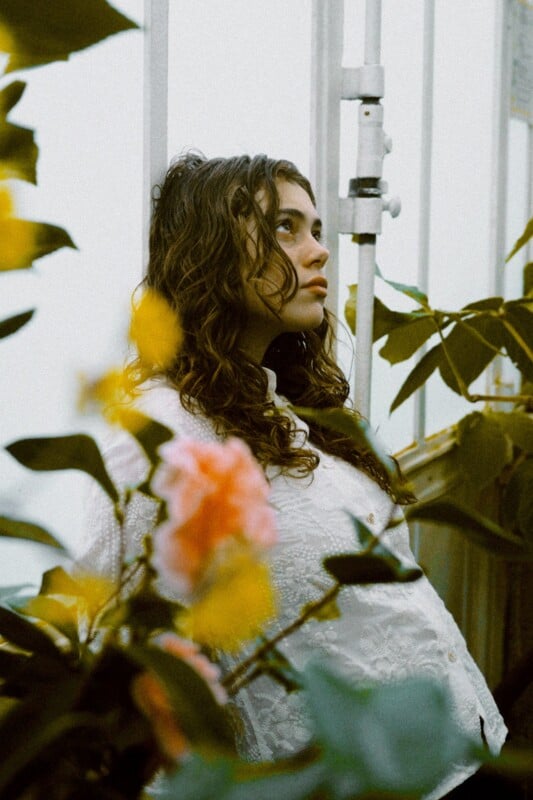 A young woman with wavy hair and a white textured top leans against a greenhouse window, surrounded by green foliage and soft yellow flowers, gazing thoughtfully upward.