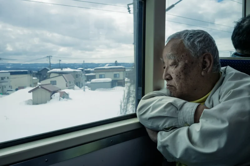 An elderly person with gray hair sits by a train window, resting their arms and head on the window ledge, gazing out at a snowy landscape with houses under a cloudy sky.