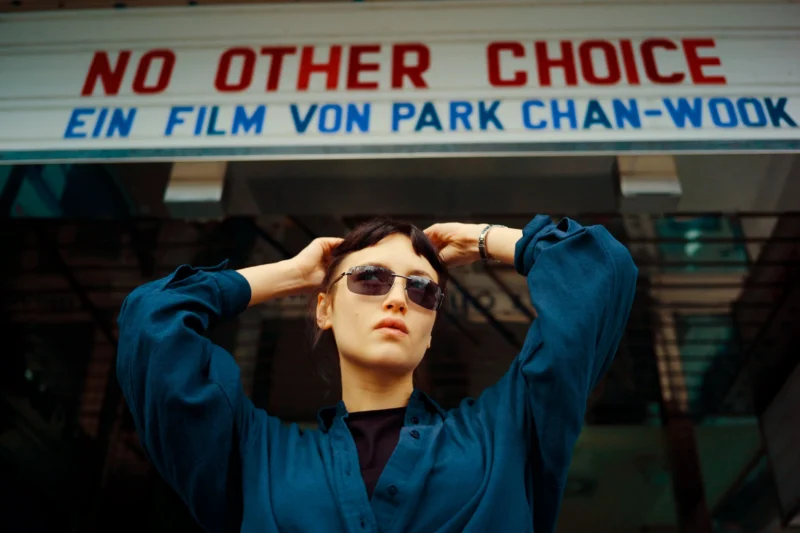A person wearing sunglasses and a blue shirt stands with hands on their head in front of a sign that reads “NO OTHER CHOICE EIN FILM VON PARK CHAN-WOOK.”.