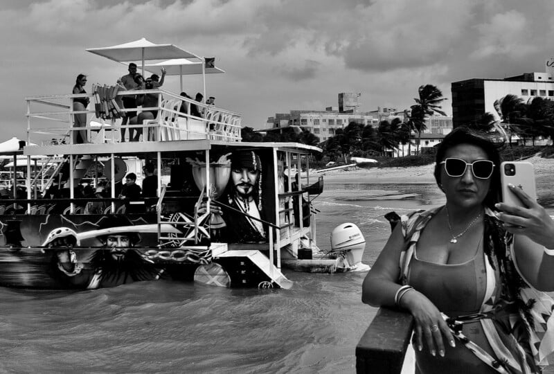 A woman wearing sunglasses takes a selfie by the shore, while a decorated boat with pirate-themed artwork and people on its upper deck floats nearby under a cloudy sky.