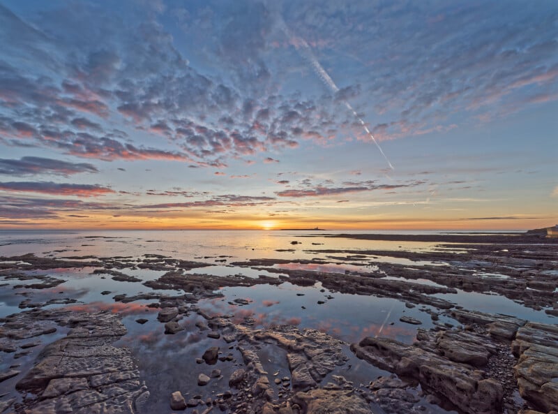 Rocky shoreline with shallow water pools reflecting a colorful sunset sky filled with clouds; the sun is low on the horizon, casting warm light across the tranquil sea and wet stones.