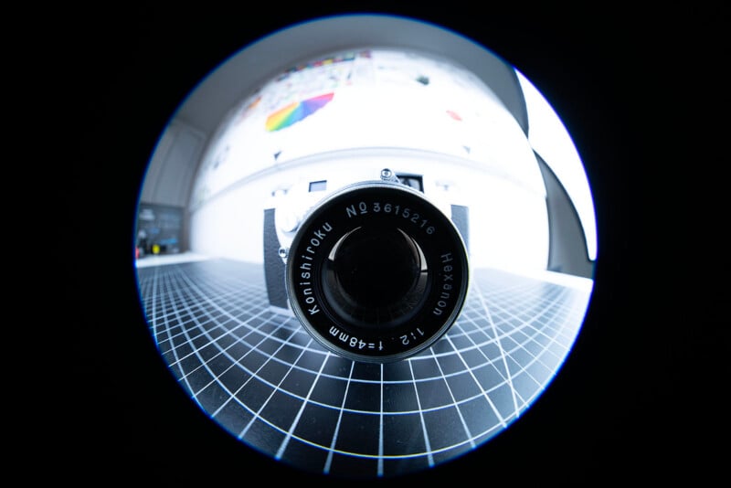 A fisheye lens photo of a camera positioned on a grid-patterned surface, with the camera lens marked "Konishiroku Hexanon 1:2 f=48mm" at the center, and a blurred background with colorful elements.