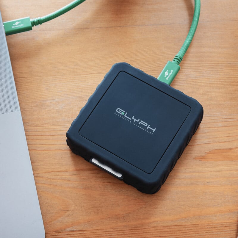 A black Glyph external hard drive with a rugged case is connected to a laptop via a green USB cable, all placed on a wooden surface.