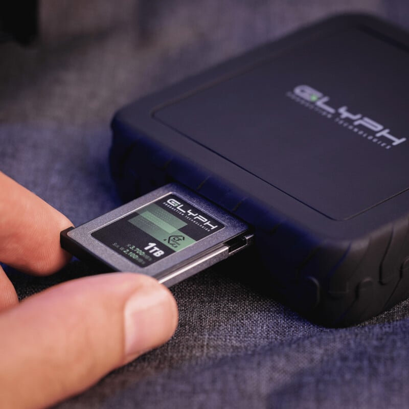 A hand inserts a Glyph 1TB memory card into a black Glyph portable storage device on a dark fabric surface.
