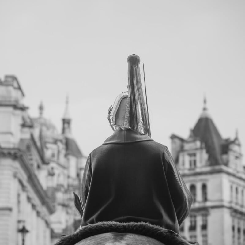 A person in ceremonial uniform and helmet sits on horseback, facing away, with historic European-style buildings in the background. The image is in black and white.