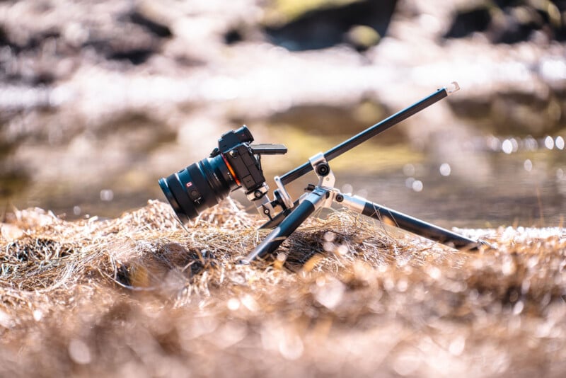 A DSLR camera with a large lens is mounted on a low-angle tripod, positioned on dry grass near a body of water, with a blurred natural background.