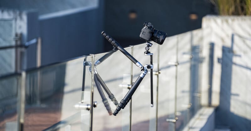 A camera mounted on a tripod is balanced precariously on a glass railing, with two legs of the tripod resting on one side and the third leg extended to the opposite side for support.