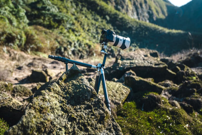 A camera with a large zoom lens is mounted on a tripod and positioned among mossy rocks in a rugged, sunlit outdoor landscape with green hills in the background.