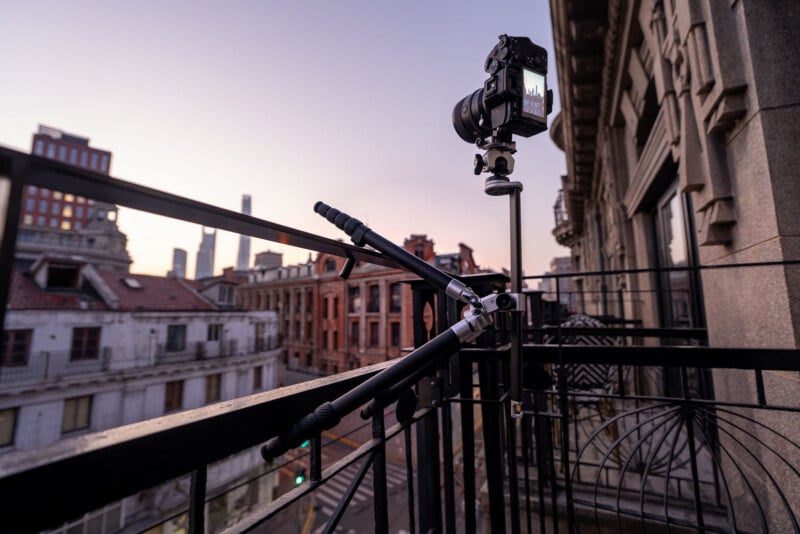 A camera mounted on a tripod stands on a balcony railing, capturing a cityscape with old and modern buildings at sunset.