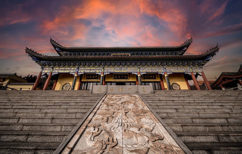 A traditional Chinese temple with ornate roof and decorated entrance, viewed from the base of wide stone steps with intricate carvings, set against a dramatic sunset sky.