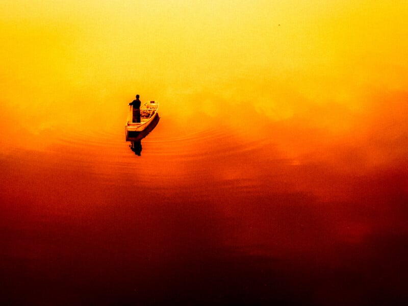 A person stands on a small boat in the middle of calm water, surrounded by vibrant yellow and orange hues, creating a surreal, dreamlike atmosphere with gentle ripples around the boat.