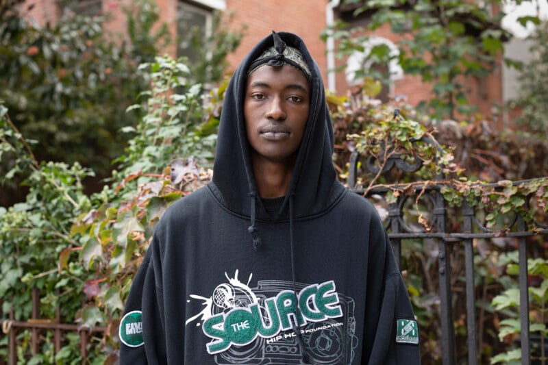A young person wearing a black hoodie with a "The Source Hip Hop Music Awards" graphic stands outdoors in front of leafy bushes and a brick building, looking directly at the camera.