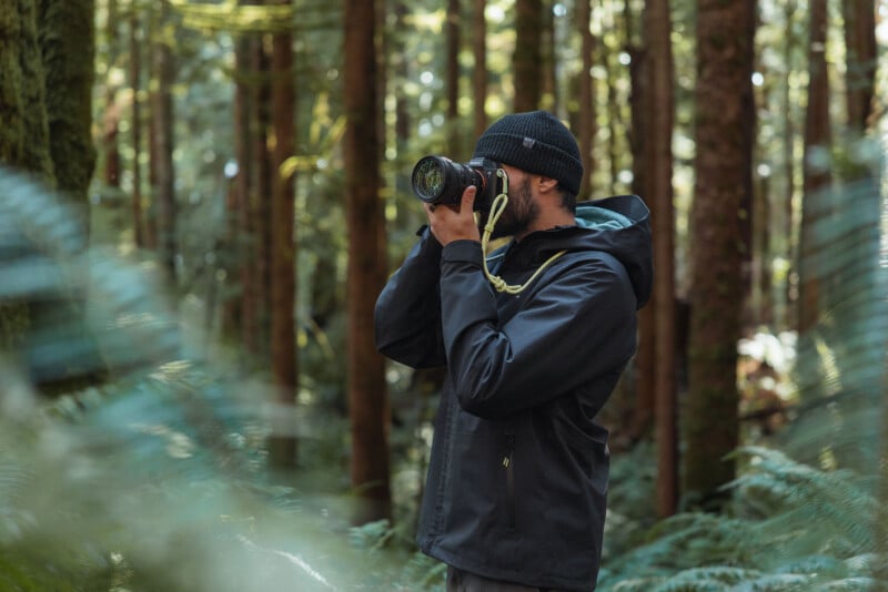 A person in a black jacket and beanie takes a photo with a camera while standing in a lush, green forest surrounded by tall trees and ferns.