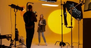 A photographer takes pictures of a woman posing in a photo studio with a yellow backdrop, professional lighting equipment, and camera gear visible around them.