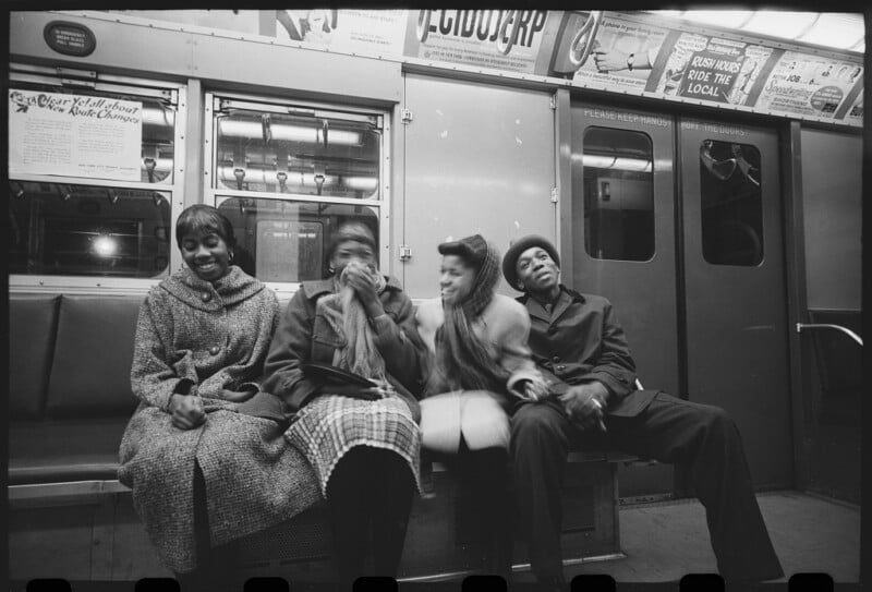 Four people sit together on a subway bench, laughing and smiling. The train interior includes handrails, notices, and advertisements above and behind them, creating a lively, candid moment.
