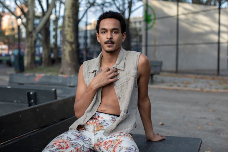 A person with short curly hair, wearing a sleeveless beige vest and floral pants, sits on an outdoor bench in a park, resting one hand on their chest and looking at the camera. Trees and a fence are visible in the background.
